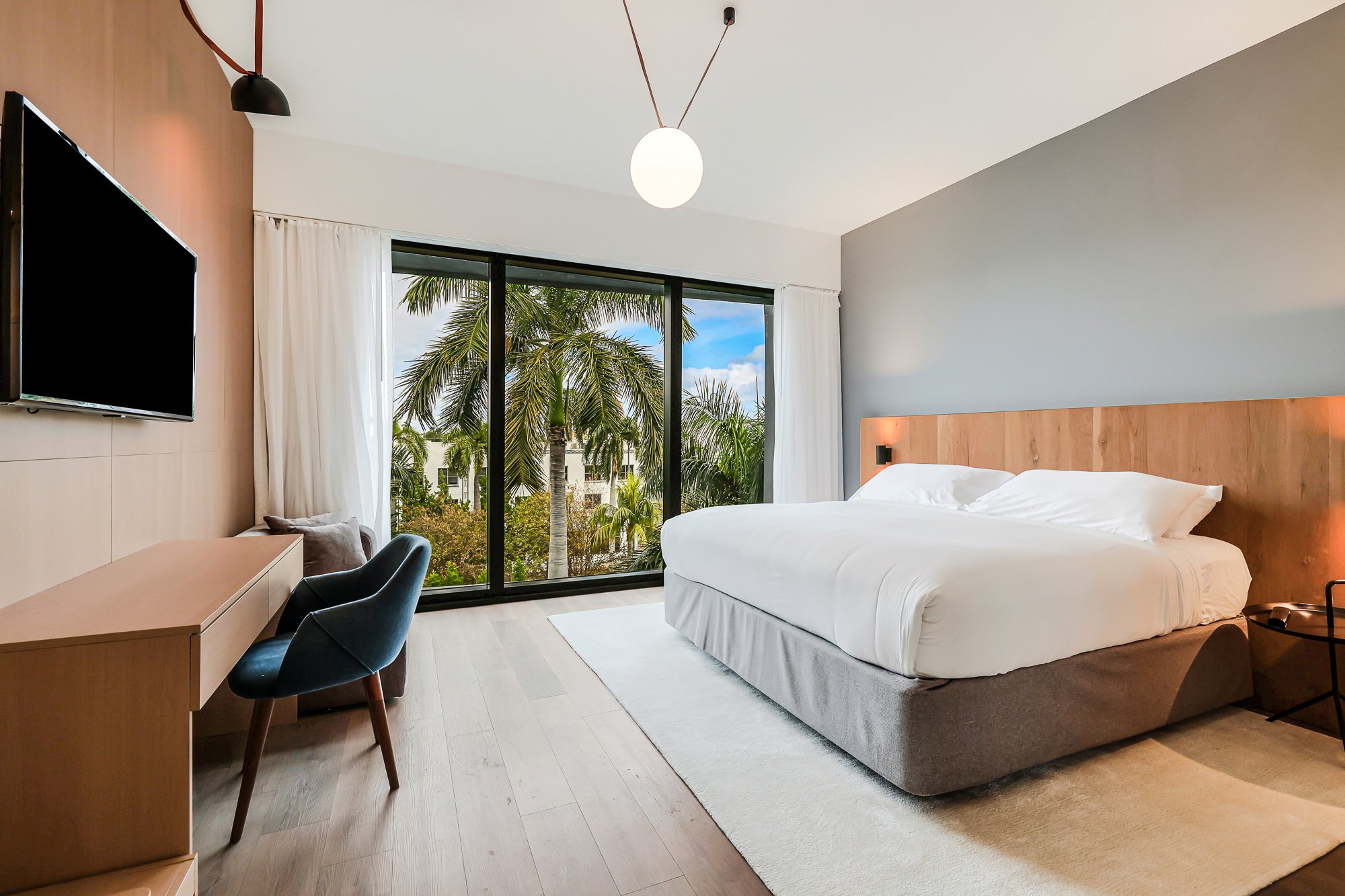 A modern hotel room featuring a large bed with white linens, a wooden desk with a blue velvet chair, and a large window showcasing palm trees outside.