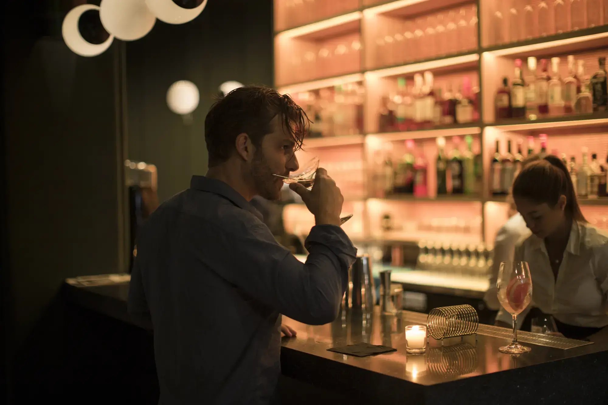 A man sipping a cocktail at an upscale, warmly lit hotel bar with glowing liquor shelves in the background.