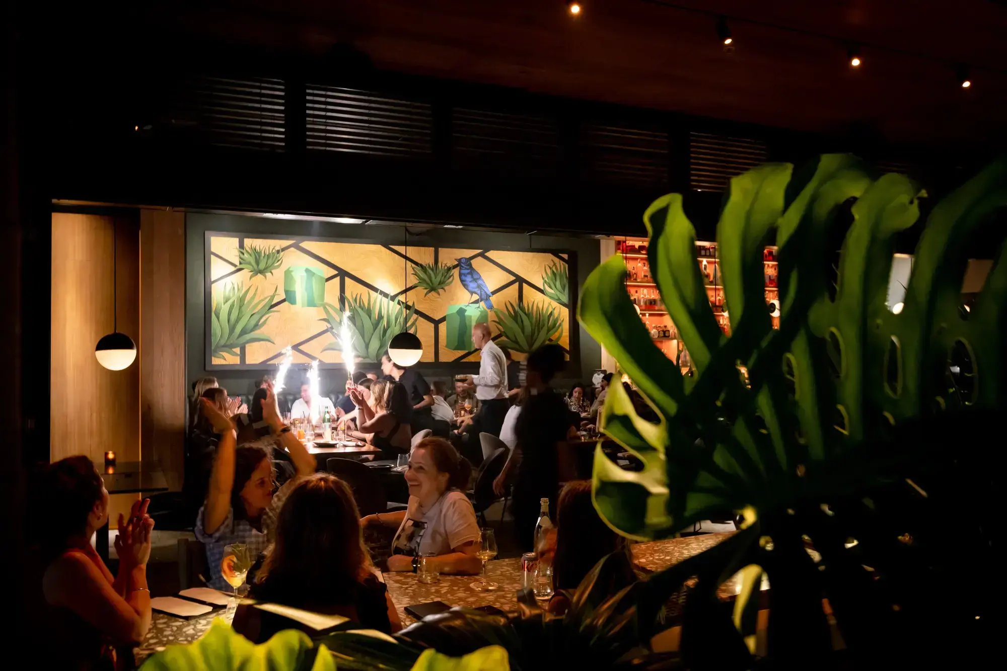 A vibrant restaurant dining room filled with celebrating guests and sparkling lights, viewed from behind a lush tropical monstera leaf.