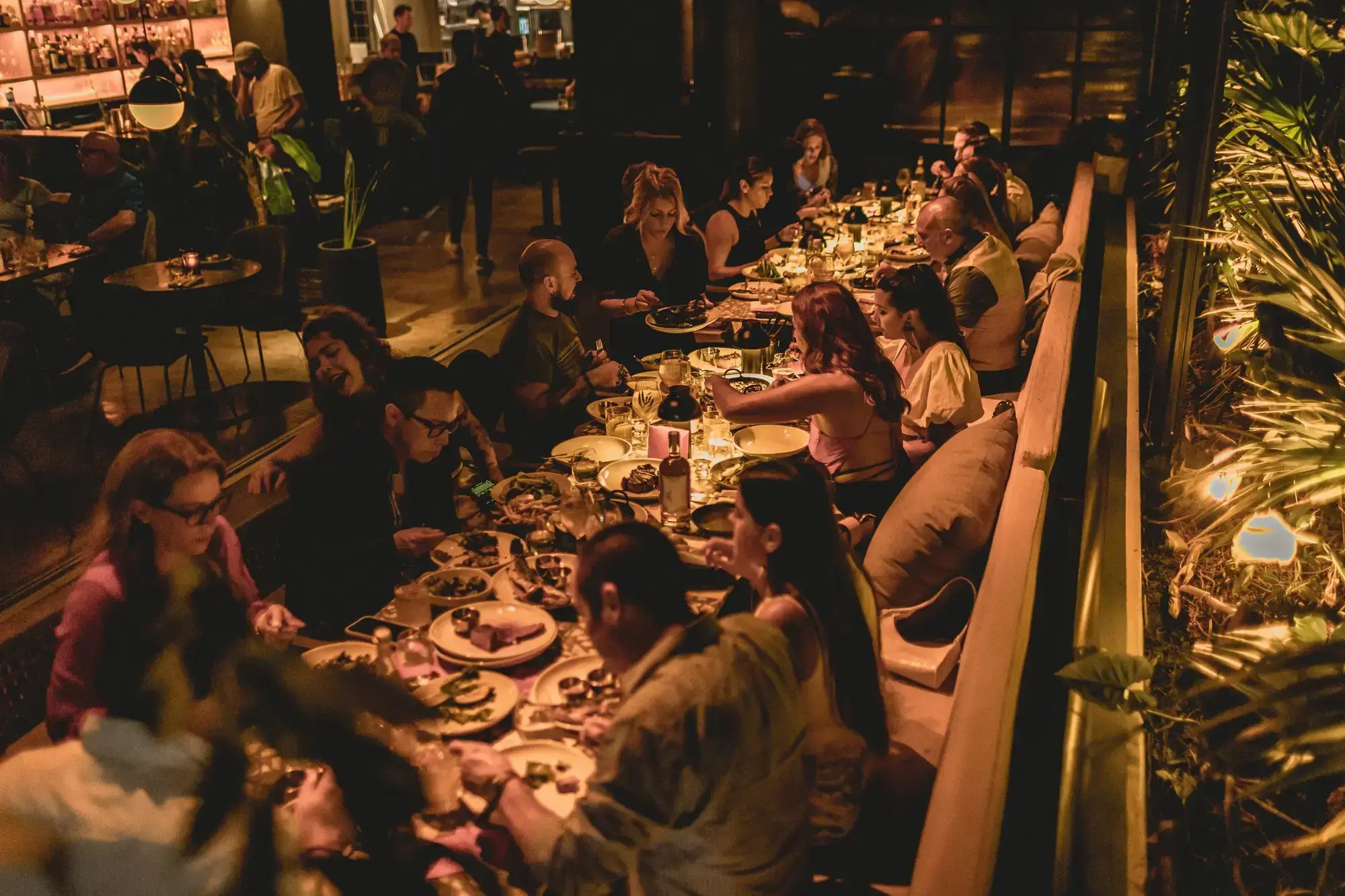 A group of people enjoying dinner and drinks along a lively, warmly lit banquet table in a restaurant patio setting.