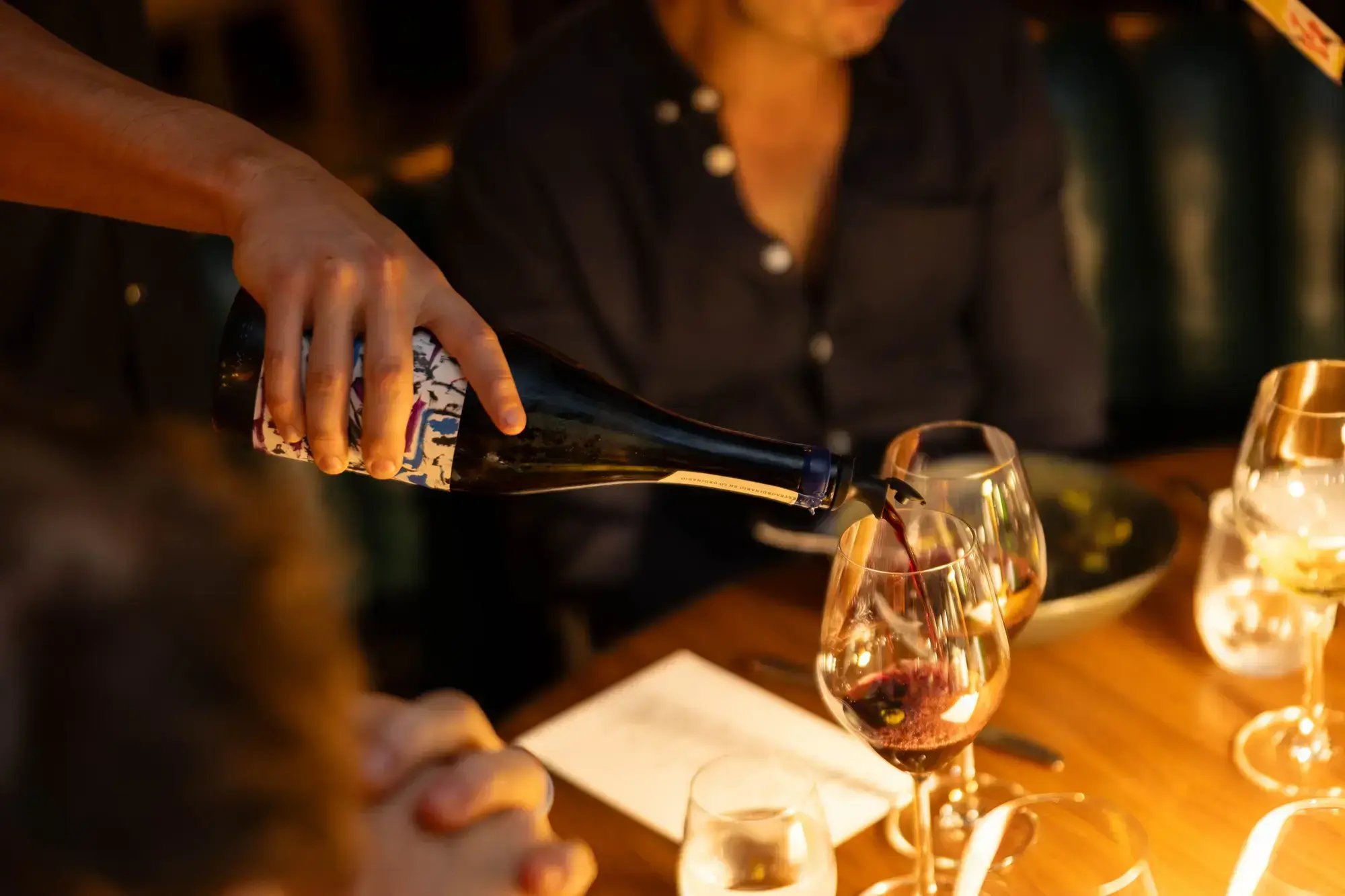 A server pouring red wine into a stemware glass at an intimate, candlelit restaurant table with seated diners.