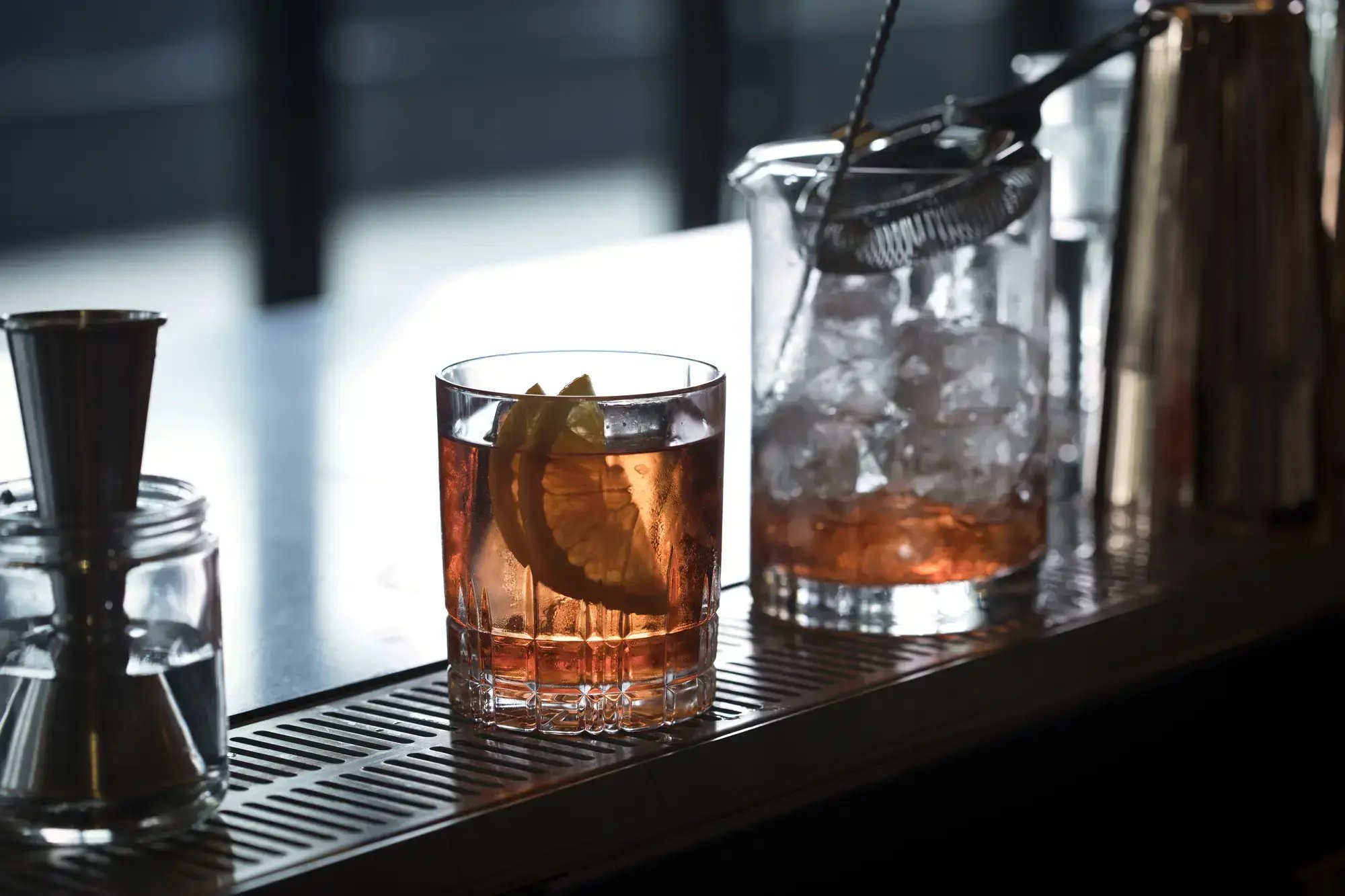 An amber cocktail garnished with an orange slice on a bar counter next to bartending tools and a glass mixing beaker filled with ice.