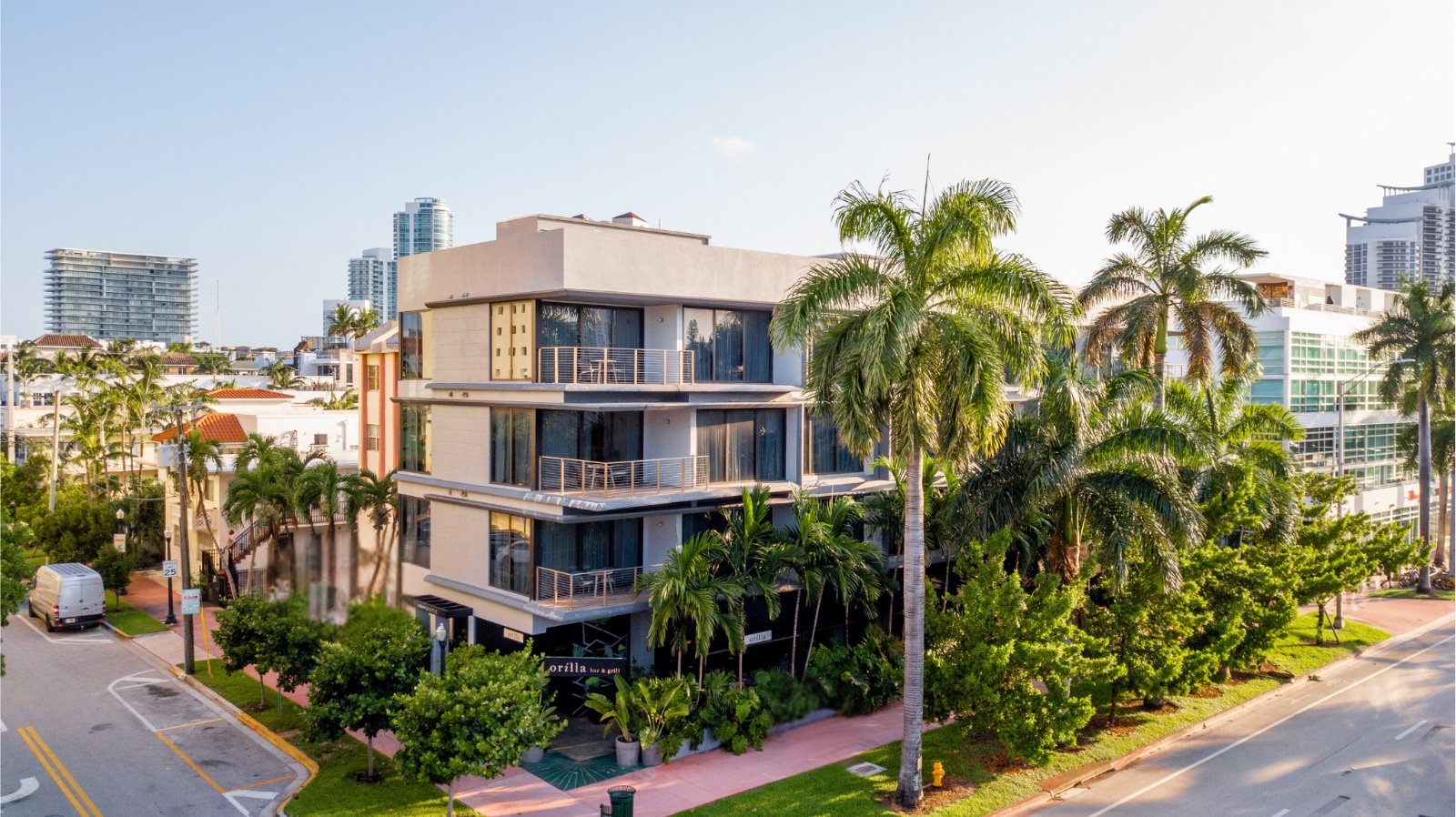 A wide exterior view of a multi-story building with balconies, surrounded by palm trees and other greenery, in a city setting.