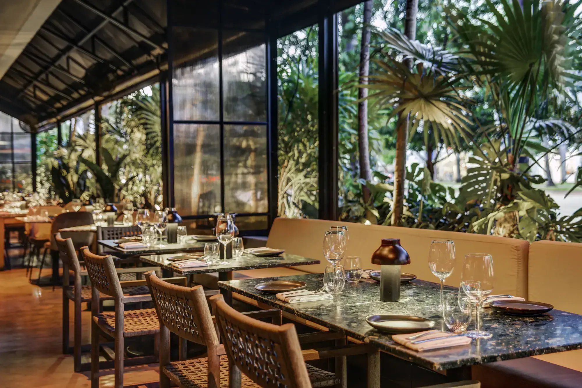 An elegant restaurant dining room with set tables alongside large windows overlooking lush tropical greenery.