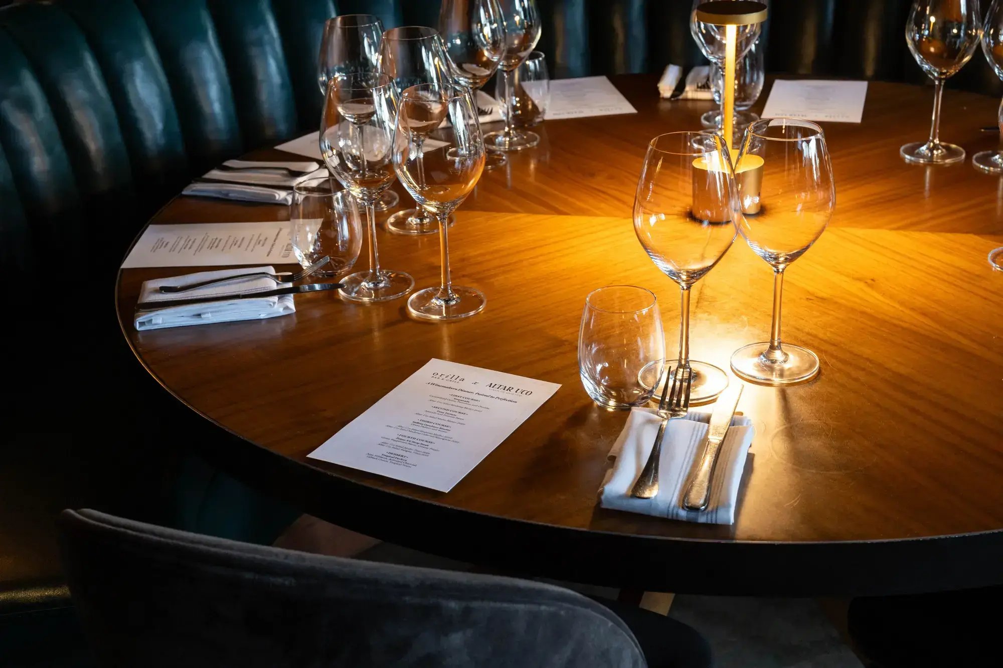 An elegantly set round wooden dining table in a restaurant, illuminated by warm candlelight with wine glasses, silverware, and printed menus.