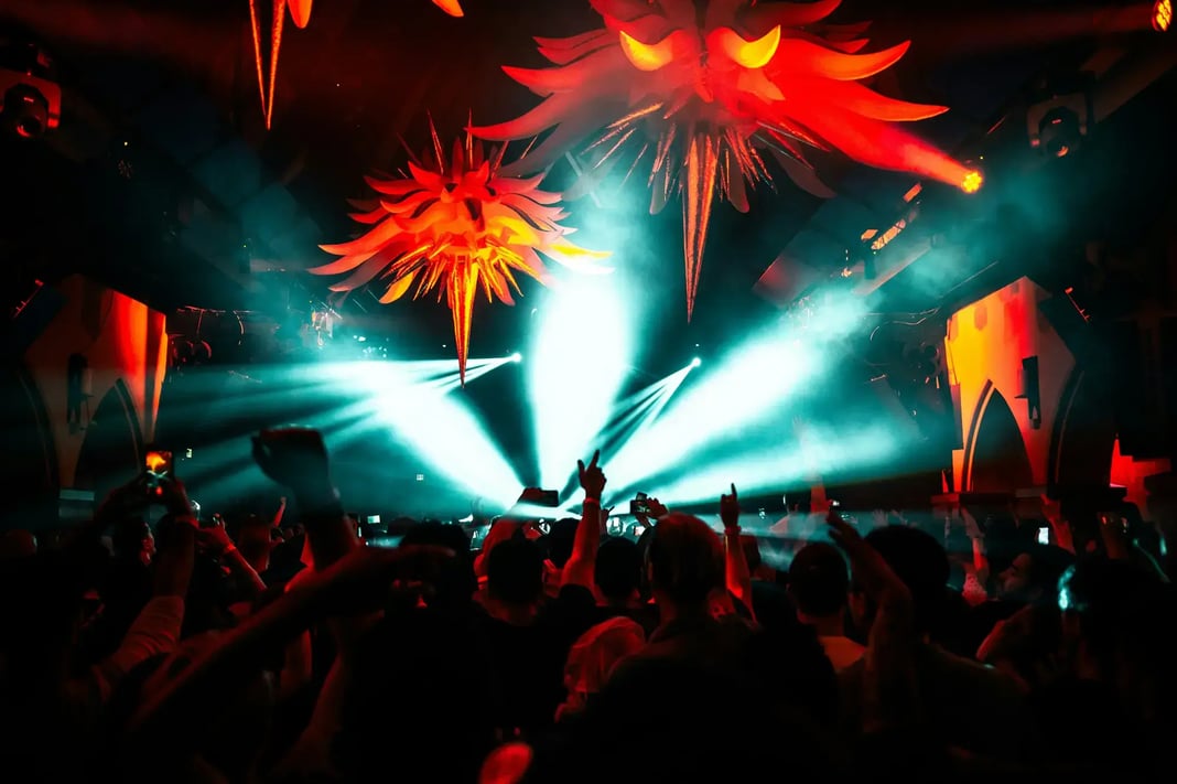 Crowded nightclub with bright stage lights, red hanging decor, and people dancing with raised hands in a high-energy nightlife scene.