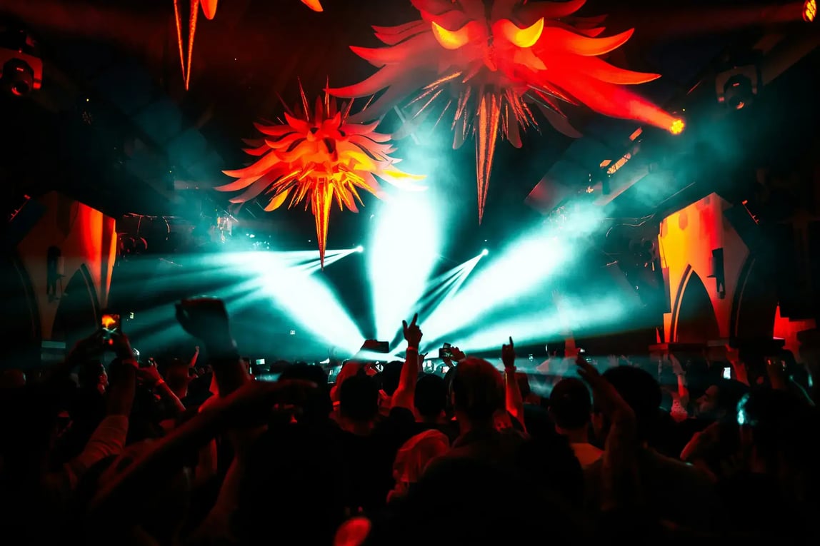 Crowded nightclub with bright stage lights, red hanging decor, and people dancing with raised hands in a high-energy nightlife scene.
