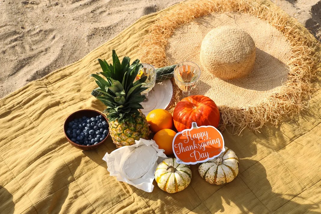 Thanksgiving-themed beach picnic setup with pumpkins, pineapple, oranges, blueberries, cheese, and a ‘Happy Thanksgiving Day’ sign on a yellow blanket and straw hat.