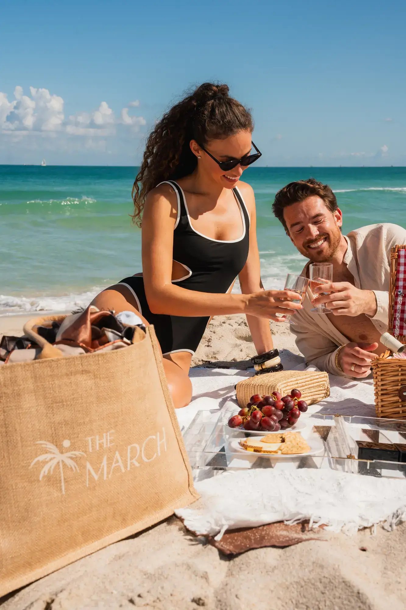 A photo of a couple picnicking on a beach, toasting with champagne next to a THE MARCH tote bag.