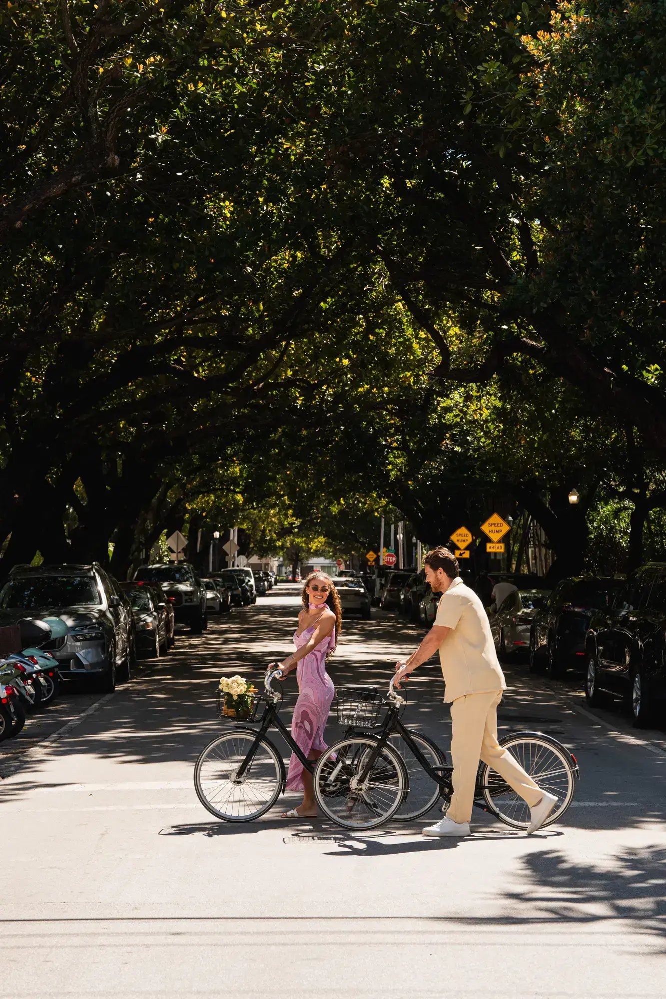 A young couple walking bicycles across a tree-lined city street on a sunny day.