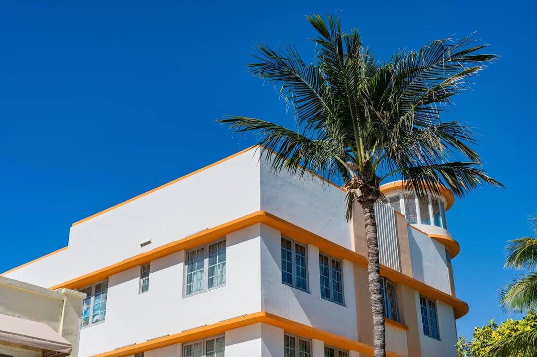 A white and orange Art Deco style building with large windows and a tall palm tree beside it, set against a bright blue sky.