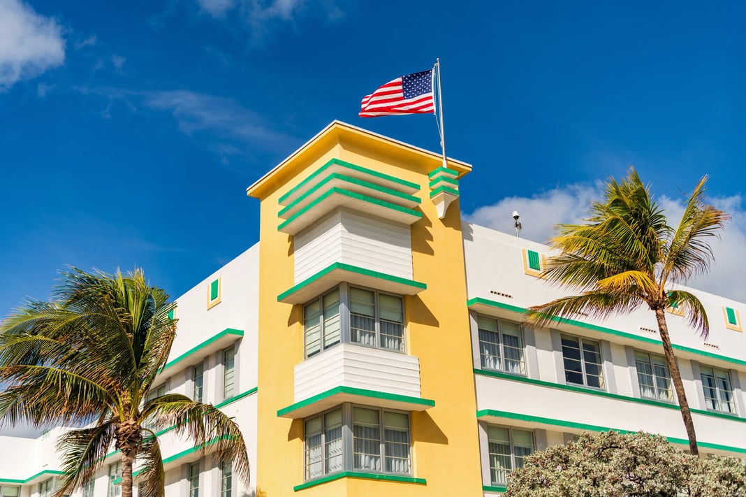 Art Deco hotel building in Miami Beach with pastel yellow and green facade, palm trees, and an American flag against a clear blue sky.